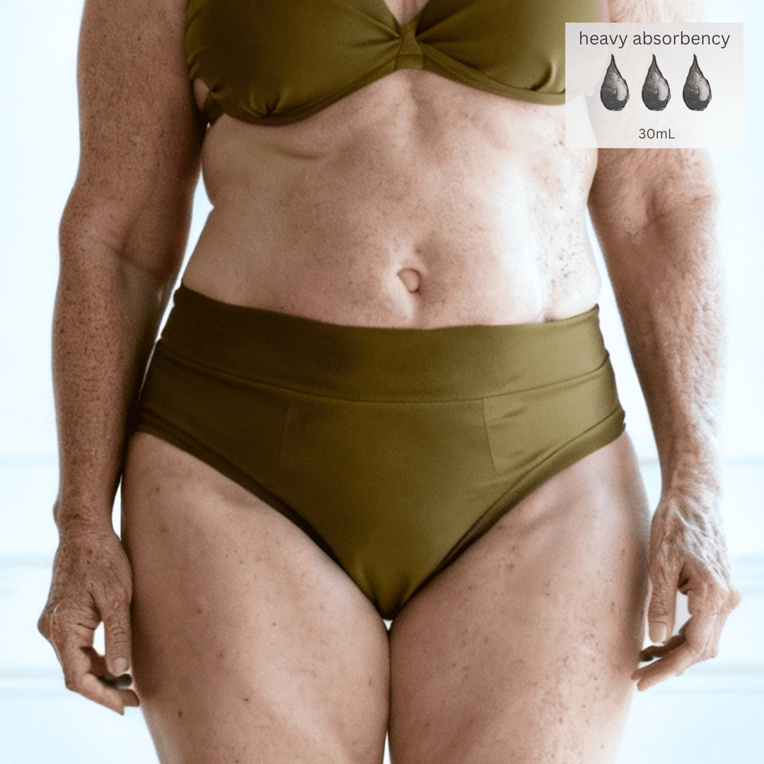 Detail of woman in her 70s wearing the High Waisted Basic brief in Moss green made from GOTS certified organic cotton. The Heavy Absorbency icon is in the top right of the image indicating this garment can absorb up to 30mL of liquid